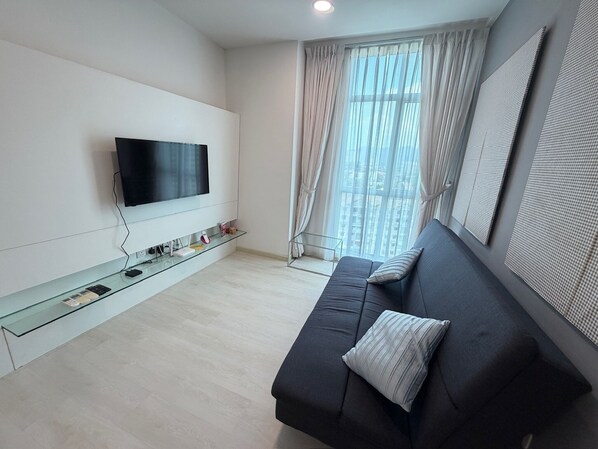 Ein modernes Wohnzimmer mit Fernseher, einem Glastisch, einer dunklen Couch mit Kissen und einem großen Fenster mit transparenten Vorhängen.