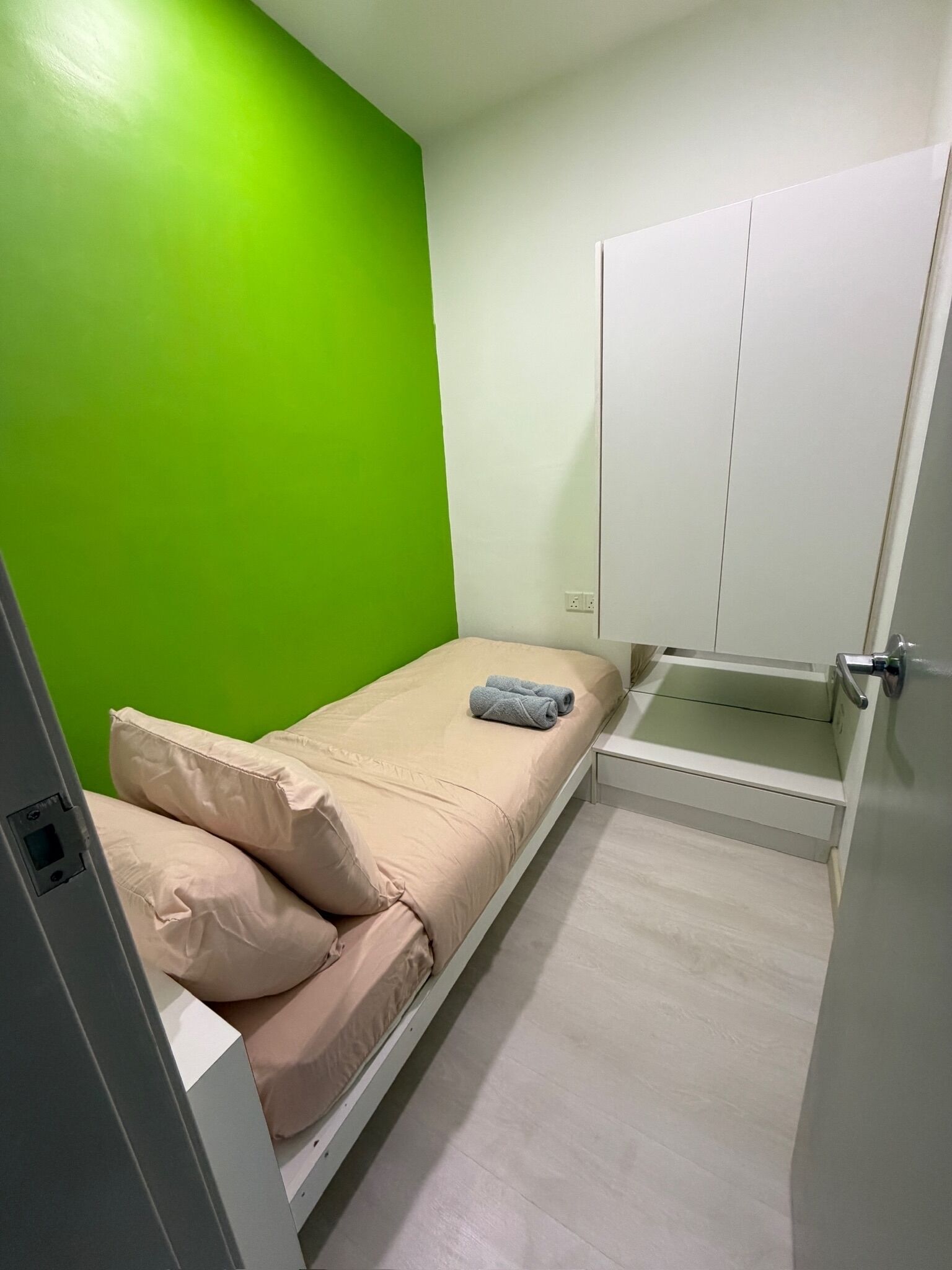 A small, single-bed room with a green wall, a white wardrobe, and a bedside table.
