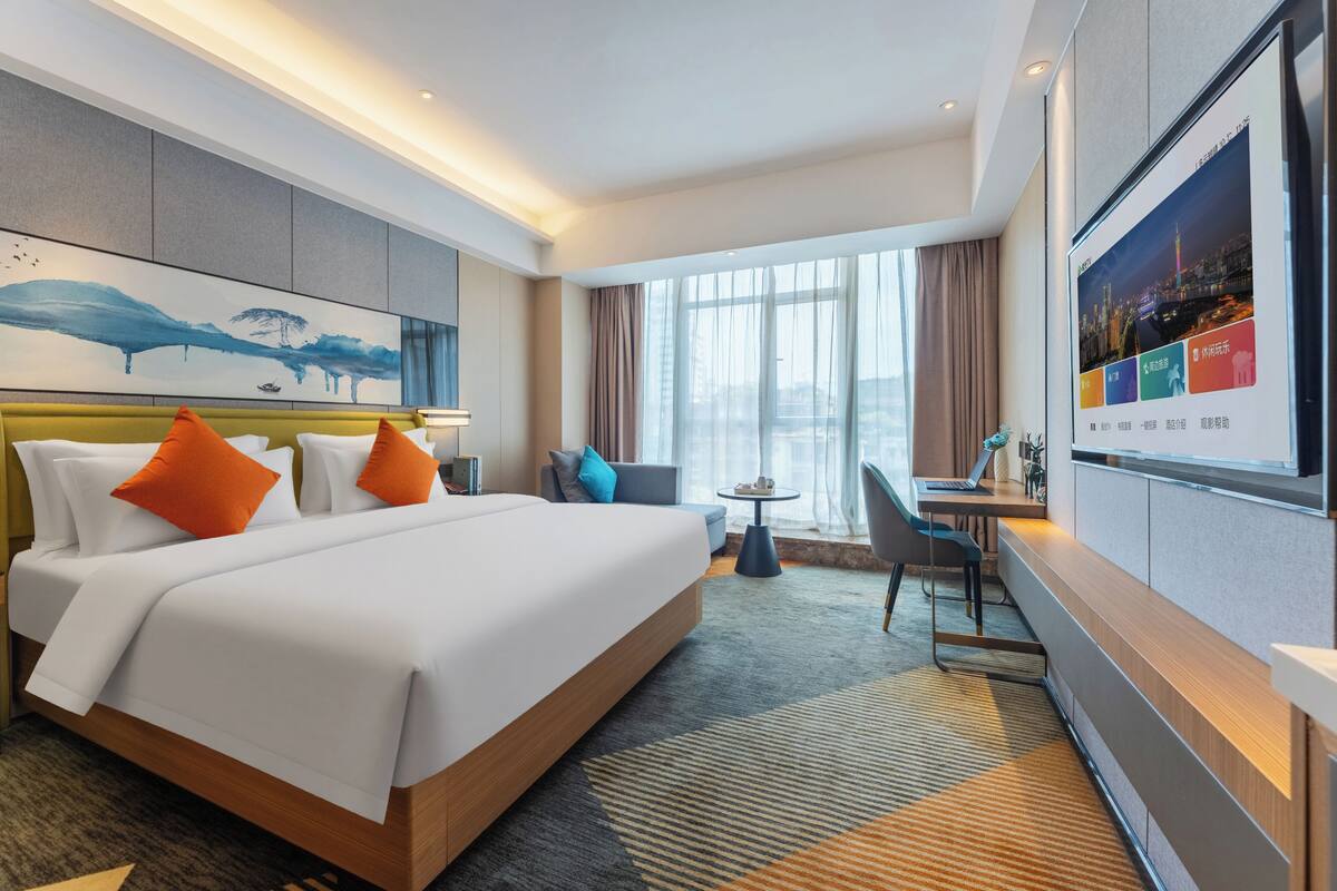 Quarto de hotel com uma cama grande, uma escrivaninha, uma cadeira, uma televisão e uma janela com cortinas.
