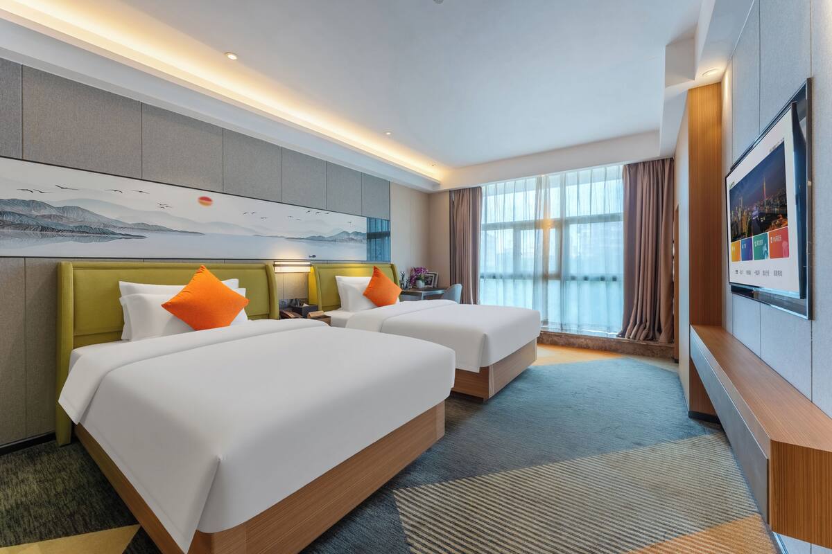 Quarto de hotel com duas camas, uma janela ampla, um quadro na parede e uma televisão de tela plana.