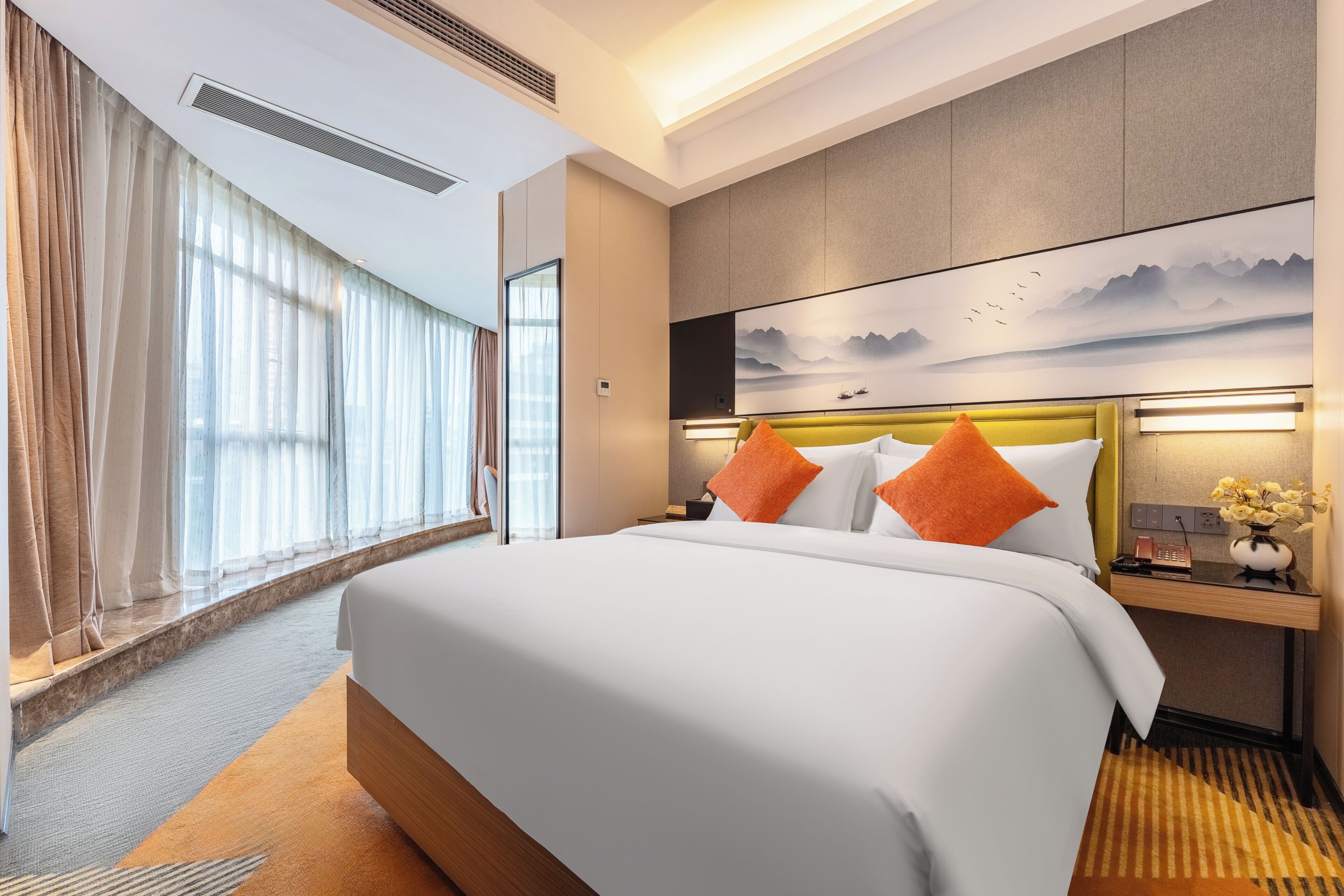 A hotel room with a large bed, a headboard with integrated lighting, a bedside table with a vase of flowers, and a view of mountains through the window.