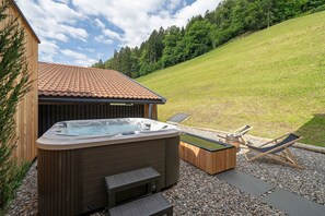 Un bain à remous avec une terrasse en bois, deux chaises longues et un bâtiment en bois doté d’un toit en tuiles.