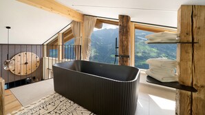 Une salle de bain moderne avec une baignoire sur pieds, des poutres en bois, une grande fenêtre donnant sur les montagnes et une horloge ronde en bois accrochée au mur.
