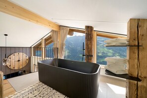 Un baño moderno con una bañera independiente, vigas de madera, un ventanal con vistas a la montaña y un reloj de madera redondo en la pared.