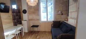 Een kamer met een houten vloer, een blauwe bank, een witte tafel met stoelen, een klein zwart tafeltje, een boekenkast, een klok en een raam met uitzicht.