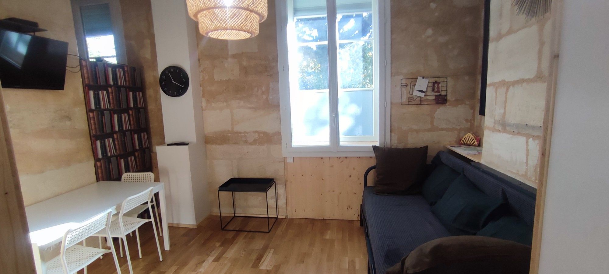 A room with a wooden floor, a white table with chairs, a bookshelf, a sofa, a clock, and a window with a view.