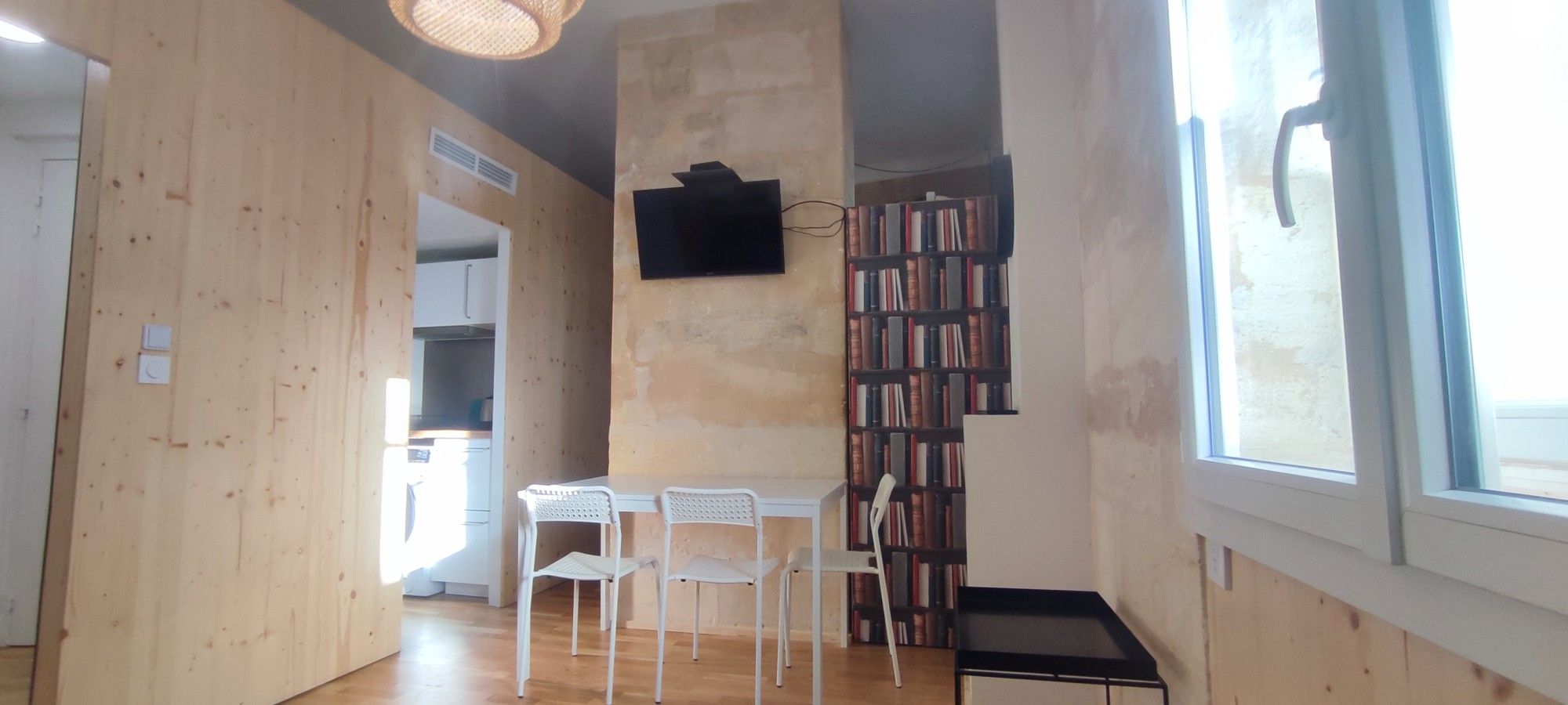 Een kamer met een houten inrichting, een eettafel met stoelen, een boekenkast en een aan de muur bevestigde televisie.
