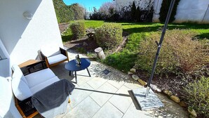 Un patio con mesa y sillas, un pequeño jardín con arbustos y césped, y un edificio con techo rojo al fondo.