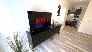 Una sala moderna con un televisor de pantalla plana mostrando 'NETFLIX', un mueble de entretenimiento negro, una planta en maceta y una cocina al fondo.