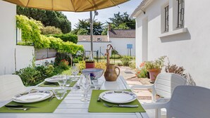 Un espace de restauration en plein air avec une table dressée pour quatre personnes, entourée de chaises blanches, sous un grand parasol.