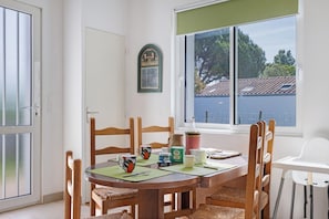 A dining area with a table set for four, wooden chairs, a white high chair, and a window with a view of a building and trees. - Spacious, Comfortable House in Barbâtre! (Barbâtre)