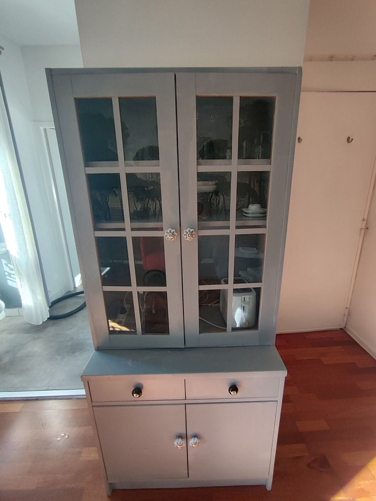 Ein weißer Schrank mit Glastüren und zwei Schubladen, der sich in einem Raum mit Holzboden und einer weißen Tür im Hintergrund befindet.