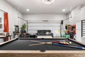 Une table de billard avec des queues et des billes, un canapé, une petite table avec une lampe, et des jeux d’arcade dans une pièce aux murs en briques blanches et dotée d’une porte de garage.
