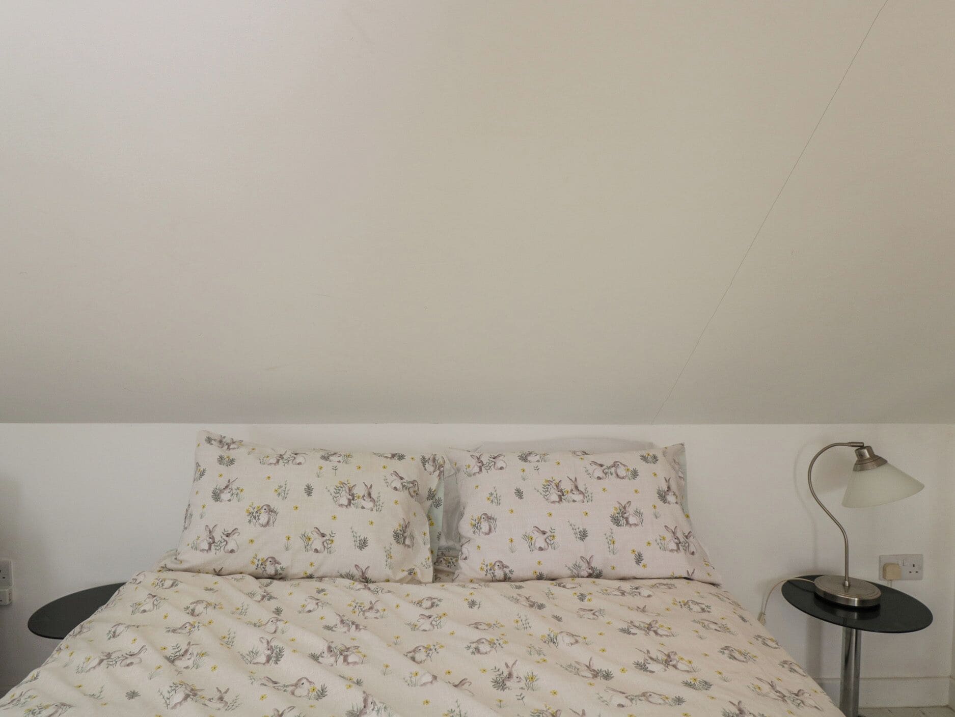 A bed with a floral print, a bedside table with a lamp, and a power outlet on the wall.