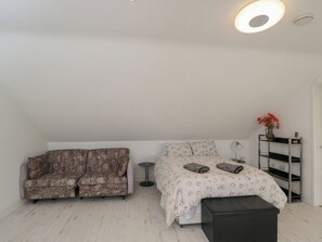 A bedroom with a bed, a sofa, a small round table, and a shelf with a vase of flowers. - The Nest (Glastonbury)