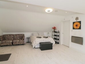 A bedroom with a sofa, a bed with a patterned bedspread, a small table, a radiator, and a wall-mounted shelf with flowers. - The Nest (Glastonbury)