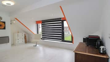 Una habitación moderna con techo inclinado, una ventana con persianas horizontales, un aparador blanco, un escritorio de madera con cafetera y un radiador en la pared.