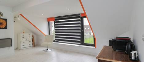 Quarto moderno com teto inclinado, janela com persianas horizontais, cômoda branca, escrivaninha de madeira com máquina de café e radiador na parede.