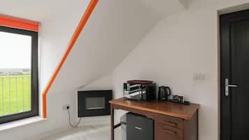 A room with a sloped ceiling, a desk with a kettle, a microwave, and a small refrigerator.