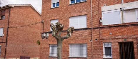 Un edificio de ladrillo de varios pisos con un árbol en frente, un banco y una entrada con portones.
