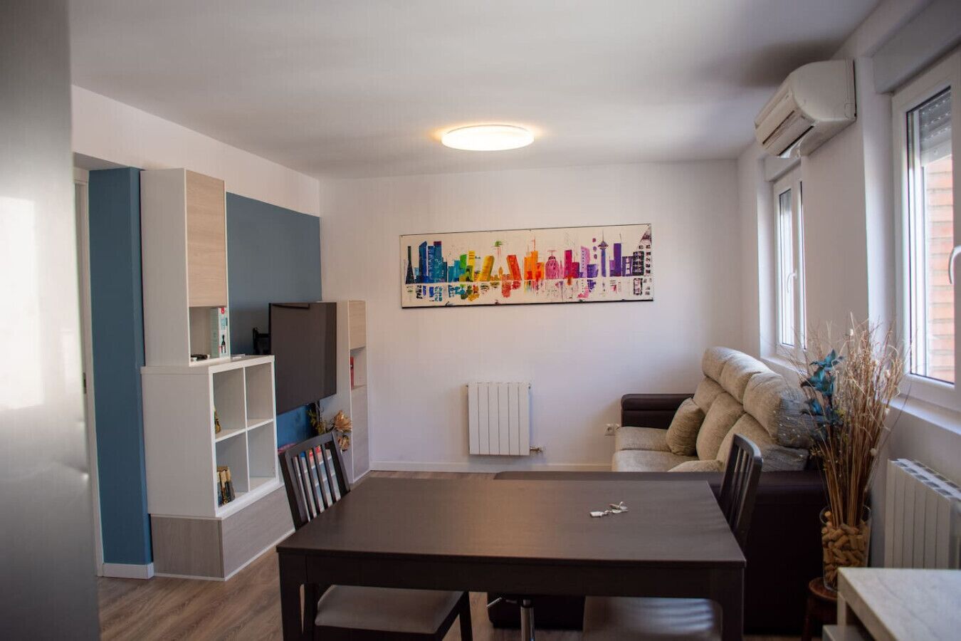 A modern living room with a dining table, a sofa, a TV, and a colorful cityscape artwork on the wall.