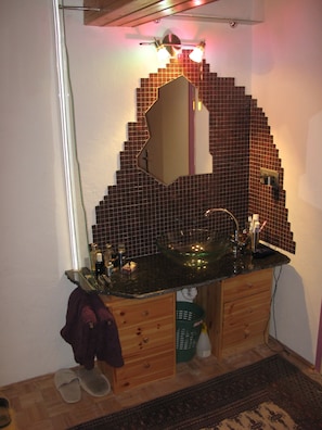 A bathroom with a tiled backsplash, a glass bowl sink, wooden cabinets, a mirror, and a green basket.