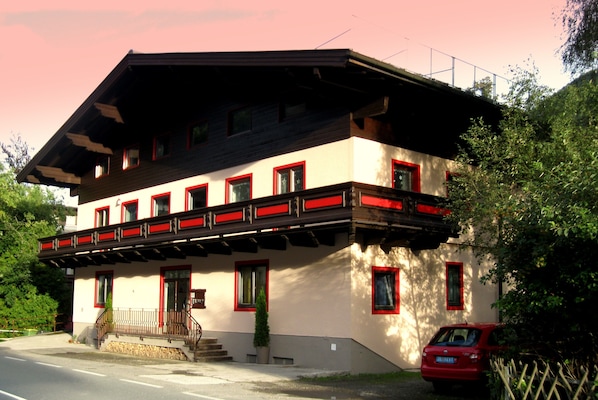A two-story building with a balcony, a red car parked in front, and a small tree in front of the entrance.