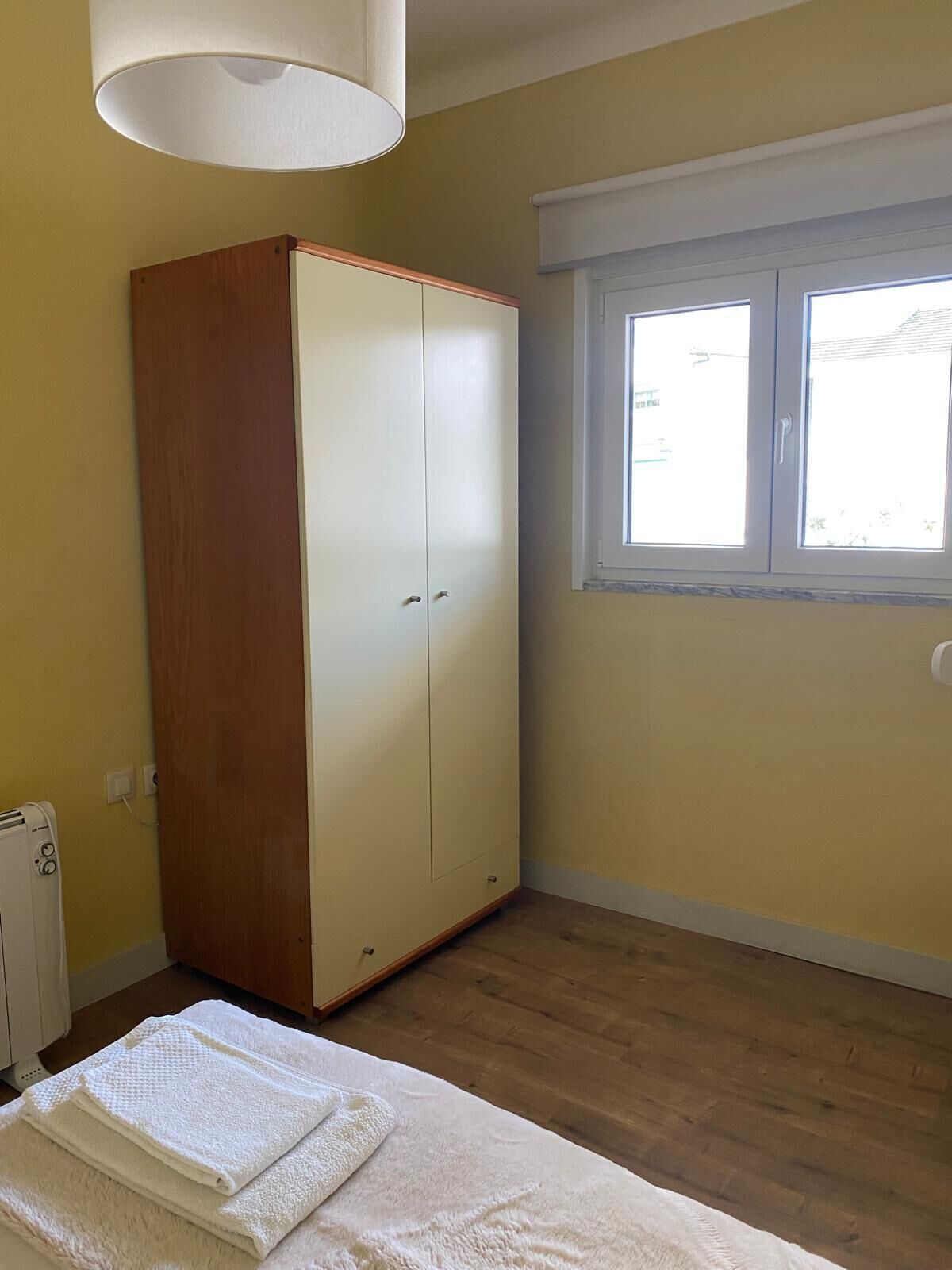 Une chambre avec une armoire, un radiateur, une fenêtre avec des stores et un lit recouvert d’une couverture blanche.