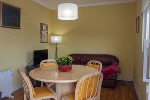 Ein Essbereich mit einem runden Tisch, Korbstühlen, einem roten Blumentopf, einem braunen Ledersofa und einer weißen Lampe.