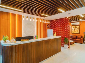 Un mostrador de recepción con diseño de panel de madera, una zona de recepción con pared de patrón rojo y un área de estar con sofá de cuero marrón.