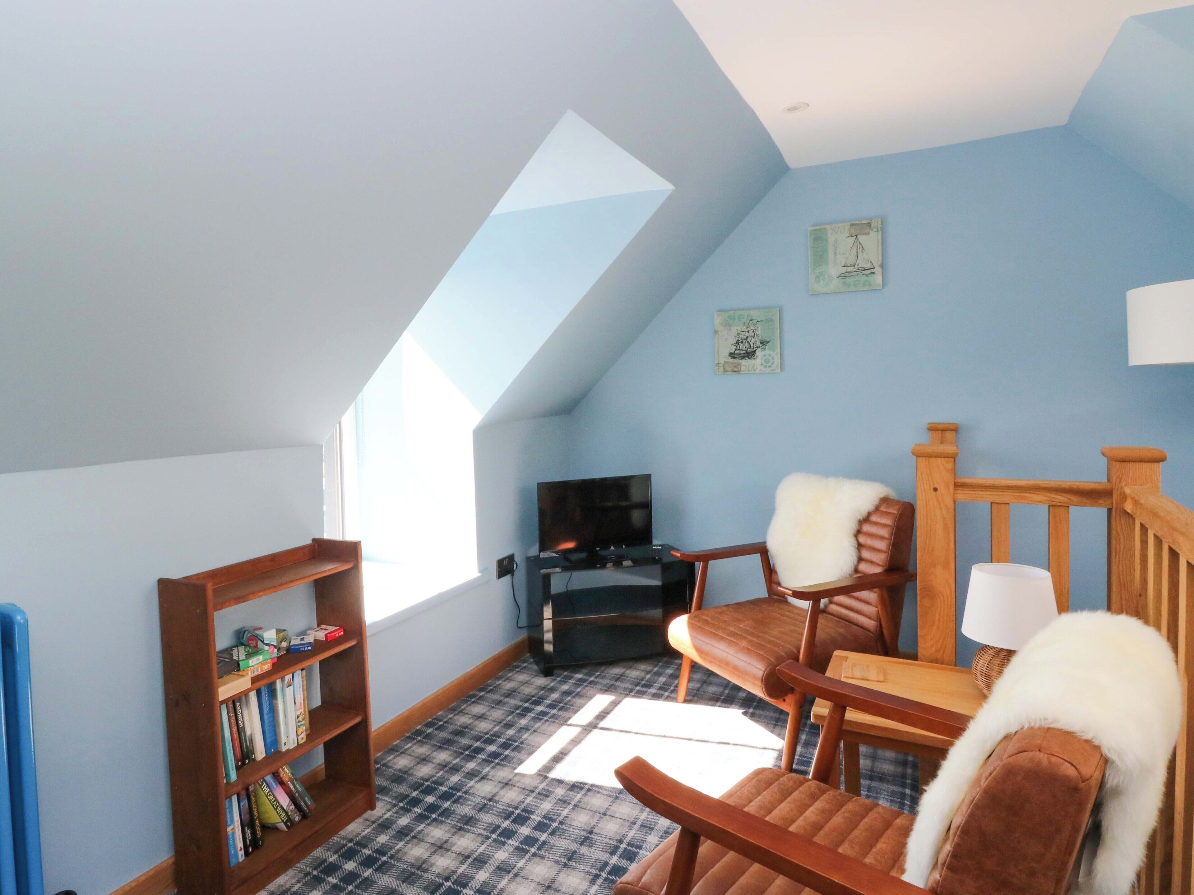 A room with a sloped ceiling, a bookshelf, a television, a brown chair with a white fur throw, and a plaid-patterned rug.