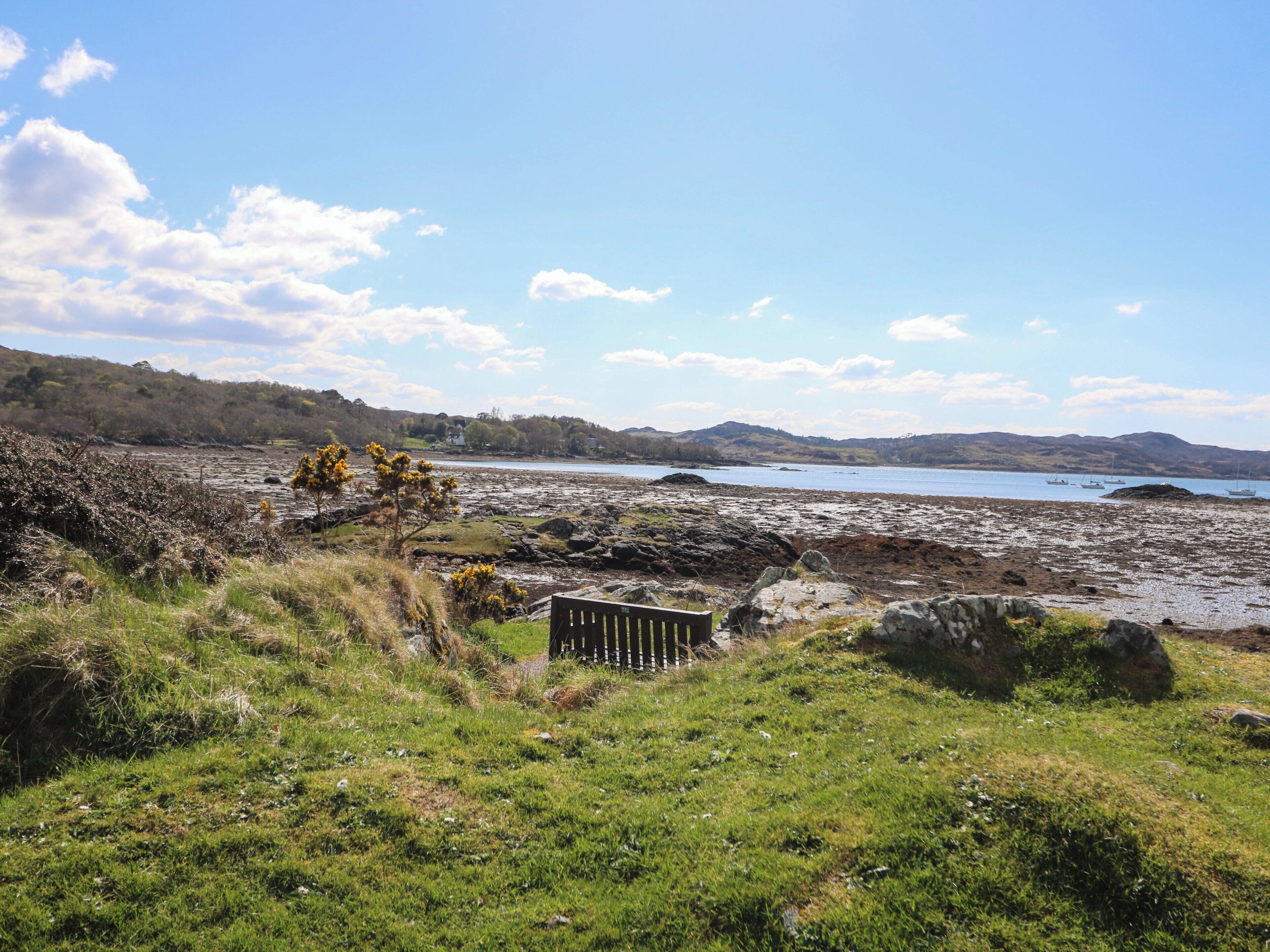 A coastal landscape with rocky terrain, a grassy foreground, and a distant view of a body of water with boats.