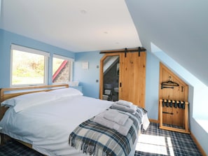 A bedroom with a bed, a window, a wooden door, and a towel rack. - Post Cottage (Arisaig)