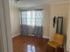 Ein Zimmer mit Holzboden, einem gelben Stuhl, einem Fenster mit gemusterten Vorhängen und einem Kleiderständer mit Kleiderbügeln.