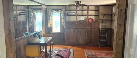 Una habitación rústica con paredes de madera, una mesa de comedor con sillas, un ventilador de techo y una pequeña cocina con una escalera.
