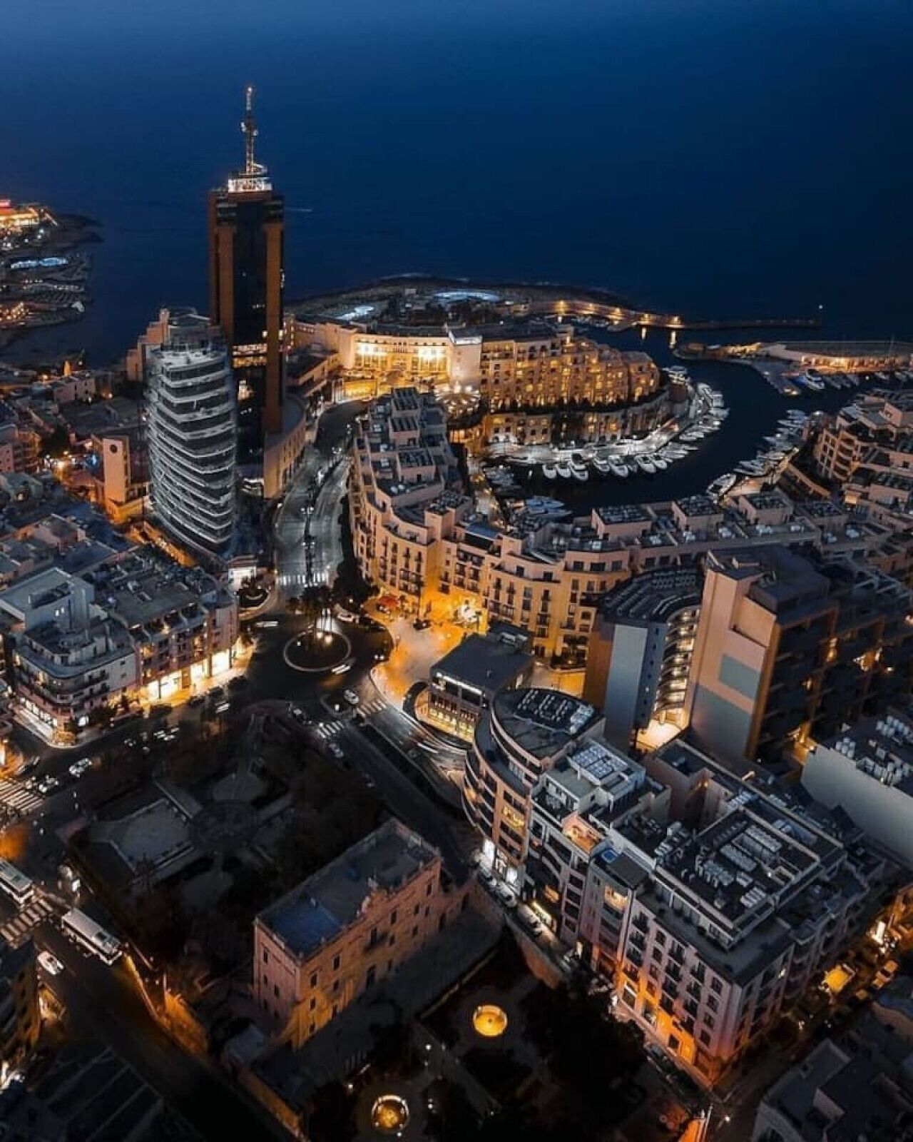 Vue aérienne d’une ville côtière de nuit, avec ses bâtiments illuminés, un port de plaisance et une tour imposante.
