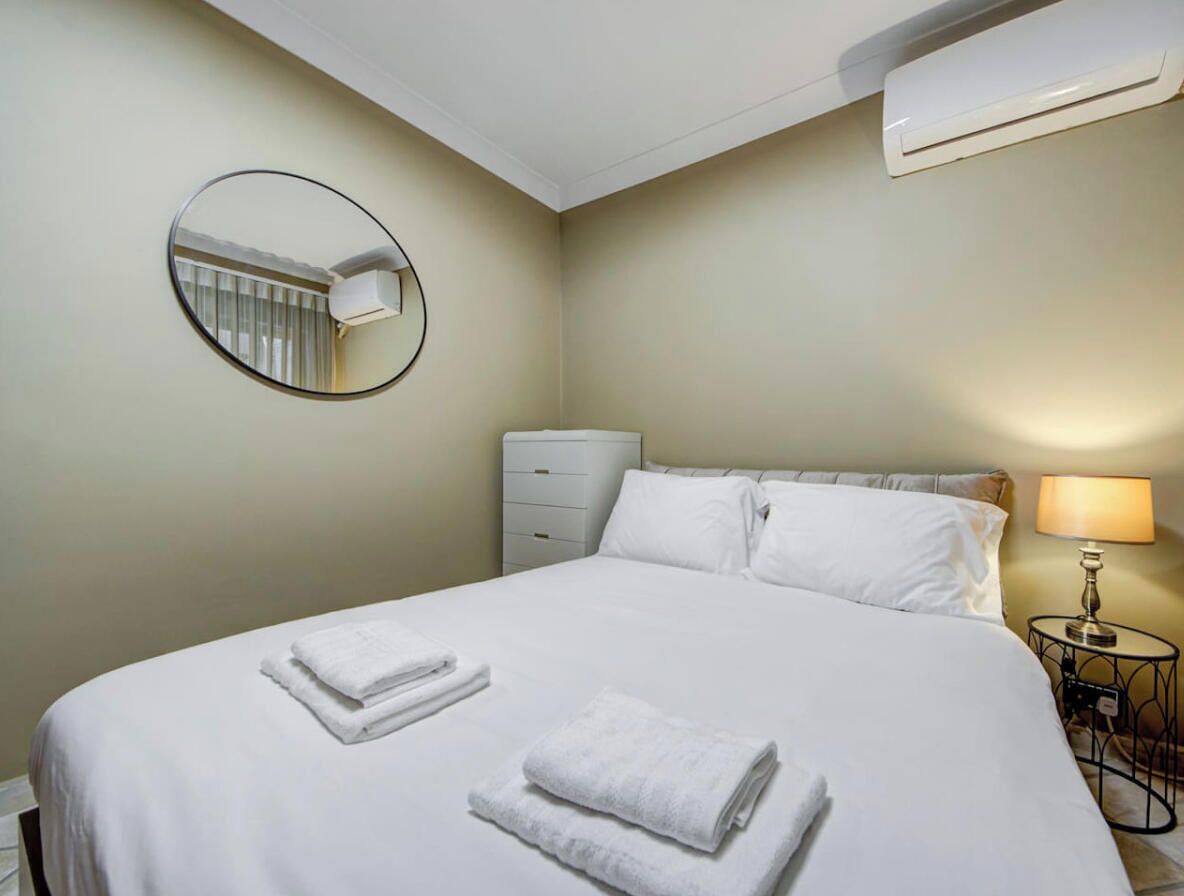 Ett hotellrum med en säng, en rund spegel, ett sängbord med en lampa och en väggmonterad luftkonditioneringsanläggning.