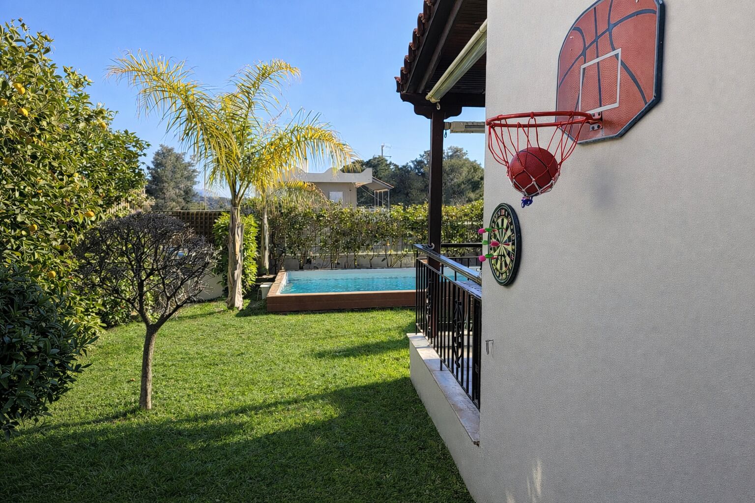 A poolside area with a basketball hoop and dartboard on a wall, surrounded by greenery and a clear blue sky.