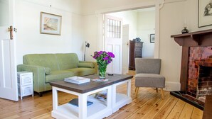 Une pièce avec un canapé vert, une table basse blanche ornée d’un vase de fleurs violettes, un fauteuil gris et une cheminée.