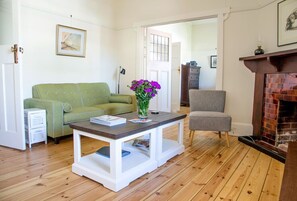 Een kamer met een groene bank, een witte salontafel met een vaas vol paarse bloemen, een grijze fauteuil en een open haard.
