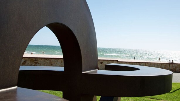 Une structure architecturale moderne au design circulaire, dotée d’un mur en pierre et offrant une vue sur la plage où l’on peut voir des gens se promener.