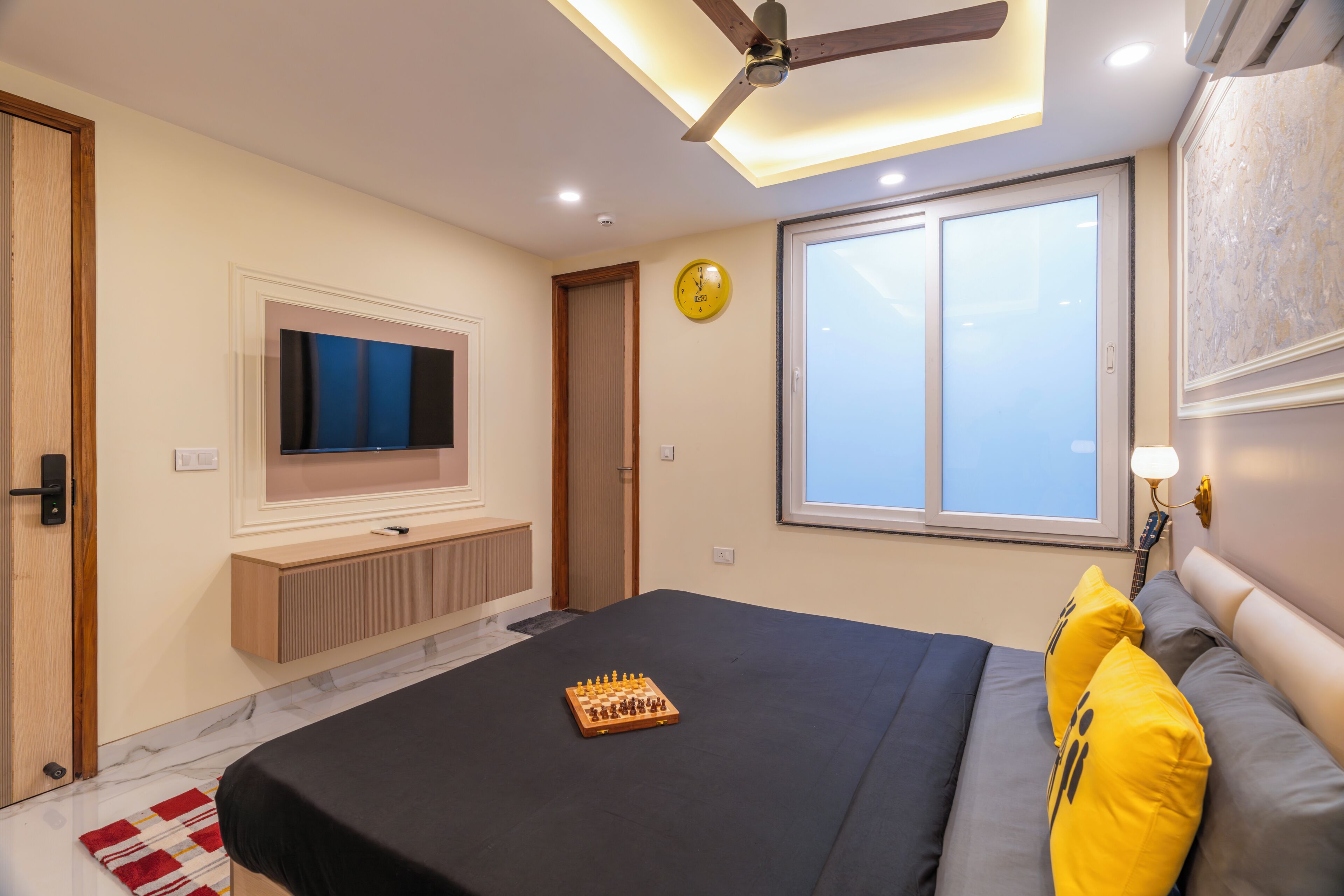 A bedroom with a bed, a ceiling fan, a mounted TV, a window, and a wall clock.