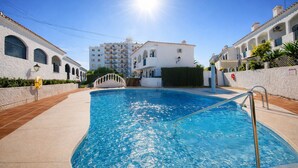 Uma piscina com água azul cristalina, rodeada por edifícios brancos com janelas em arco e varandas, e um sol brilhante no céu.