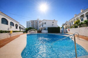 A swimming pool with a clear blue water, surrounded by white buildings with arched windows and balconies, and a bright sun in the sky. - AP426 VERANO AZUL SUNSHINE (Nerja)