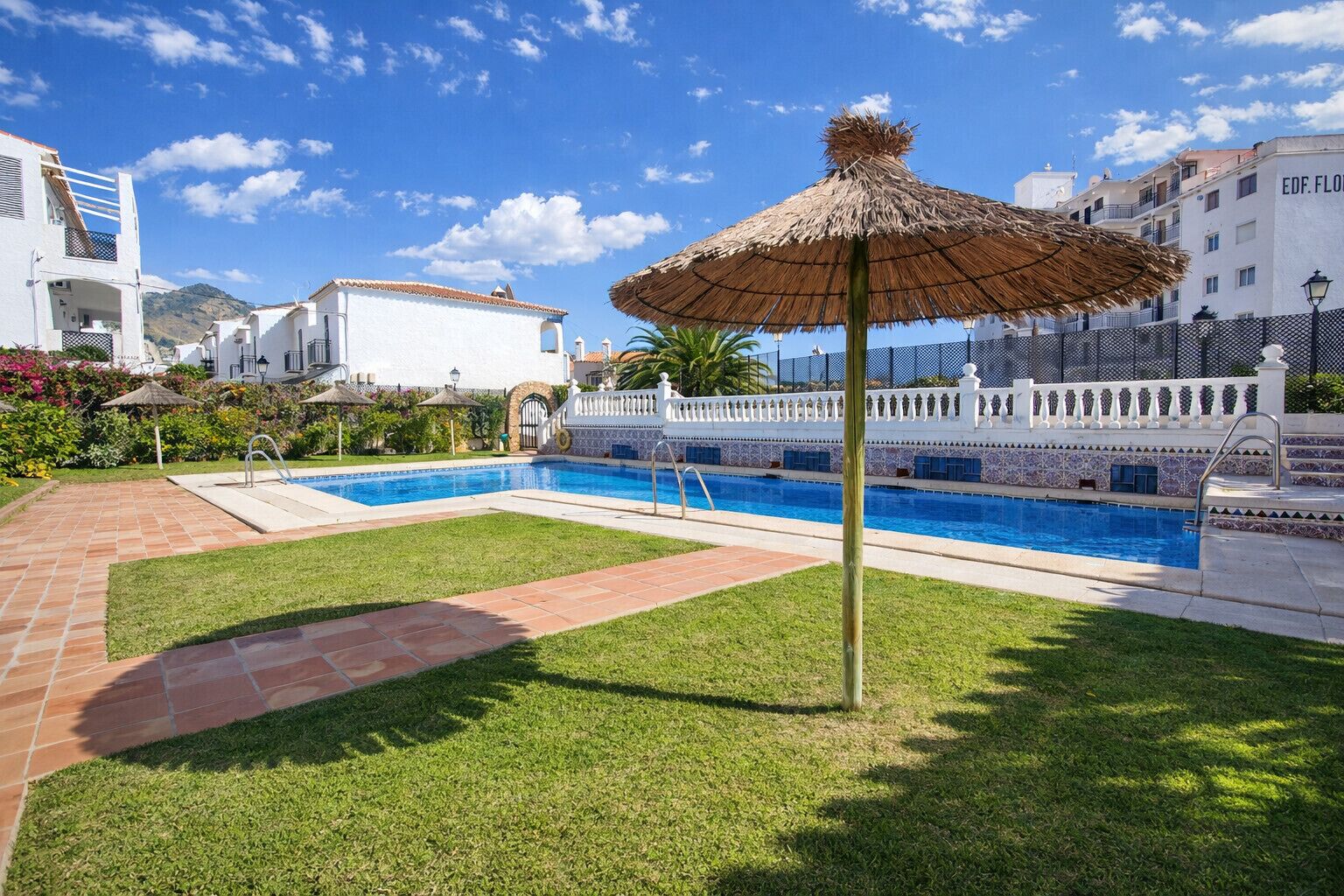 Piscina com guarda-sol de palha, cercada por uma vedação branca, e uma montanha ao fundo.