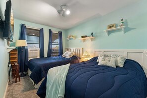 A bedroom with two beds, a ceiling fan, a window with curtains, and a shelf with decorative items.