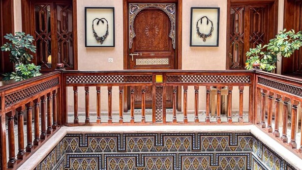 A wooden balcony with ornate railings, a patterned floor, and framed jewelry on the wall. - Riad jamaique (Marrakech)