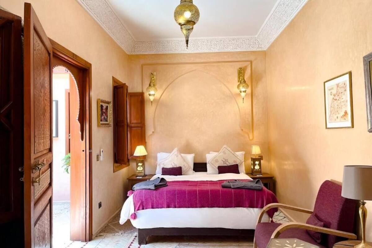 Une chambre d’hôtel avec un lit, deux lampes de chevet, un lustre, un fauteuil rouge et une petite table avec une lampe.