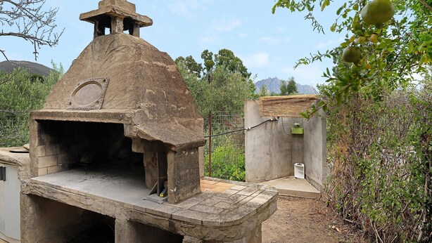 Edifici de pedra amb una xemeneia, possiblement un foc, envoltat de vegetació i un edifici petit amb teulada de rajola.