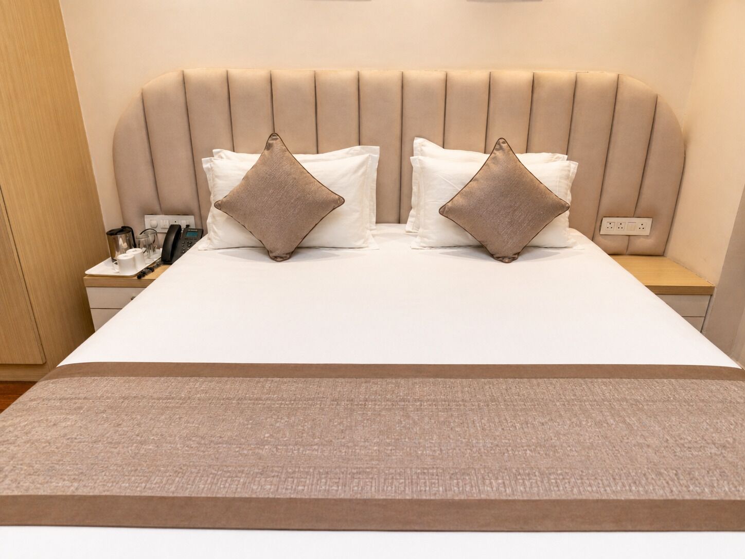 A hotel room with a bed, two bedside tables, and a nightstand with a phone and a kettle.